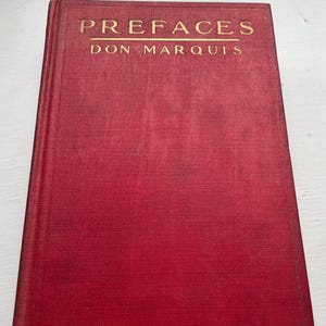Pode incluir: Um livro antigo de capa dura intitulado "PREFACES" de Don Marquis. O livro tem uma capa vermelha escura com letras douradas. A capa tem uma aparência texturizada, sugerindo uma encadernação em tecido.