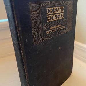 May include: A dark blue vintage book titled "Cesare Borgia" by Arthur Symons. The cover features gold lettering and decorative geometric patterns. The book shows signs of wear and age, with a slightly distressed appearance.