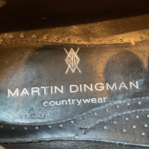 K&ouml;nnte beinhalten: Nahaufnahme der Innensohle eines schwarzen Schuhs. Der Text "MARTIN DINGMAN countrywear" ist in Silber gedruckt. Ein silbernes Logo befindet sich &uuml;ber dem Text. Die Innensohle hat einen gen&auml;hten Rand mit kleinen L&ouml;chern.