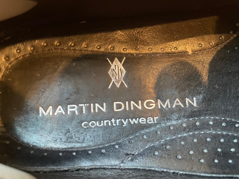 K&ouml;nnte beinhalten: Nahaufnahme der Innensohle eines schwarzen Schuhs. Der Text "MARTIN DINGMAN countrywear" ist in Silber gedruckt. Ein silbernes Logo befindet sich &uuml;ber dem Text. Die Innensohle hat einen gen&auml;hten Rand mit kleinen L&ouml;chern.