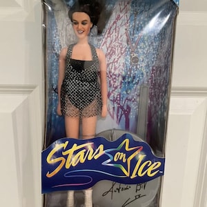 Puede incluir: Una muñeca Stars on Ice con un traje de baño de malla negra y plateada con una medalla de plata alrededor del cuello. La muñeca está empaquetada en una caja con el logotipo de Stars on Ice y la firma de un patinador artístico.