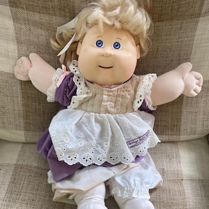 Puede incluir: Una muñeca Cabbage Patch Kids con cabello rubio, ojos azules y un vestido morado y blanco. La muñeca está sentada sobre una tela a cuadros marrón y blanco.