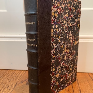Pode incluir: Livro antigo com lombada de couro escuro e capa marmorizada. A lombada é preta com o nome do autor, Prévost, e o título, La Fausse Bourgeoise, em letras douradas. A capa apresenta um fundo preto com marmoreio vermelho, dourado e branco.