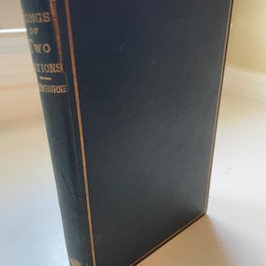 Può includere: Un libro d'epoca intitolato "Songs of Two Nations" di Swinburne. Il libro ha una copertina blu scuro con scritte e bordi dorati. Il dorso mostra usura e il libro è in piedi su una superficie bianca.