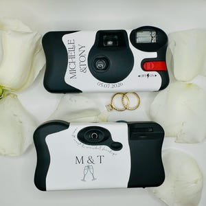 May include: Two disposable cameras with black and white casings. The top camera features the text MICHELLE & TONY and 05.07.2026. The bottom camera displays M & T with a champagne glass graphic and text. Two gold rings are positioned between the cameras.