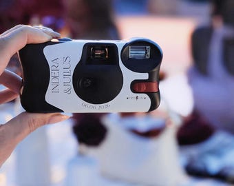May include: A disposable camera with a white and black exterior featuring the printed text INDERA & JULIUS and the date 06.06.2026. The device includes a central lens, a rectangular flash unit, and a red sliding switch labeled OFF and ON.