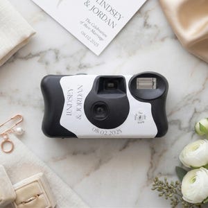 May include: A black and white disposable camera with the names "Lindsey & Jordan" and the date "08.02.2025" printed on it. The camera is surrounded by wedding-themed items, including a floral arrangement and a wedding invitation.