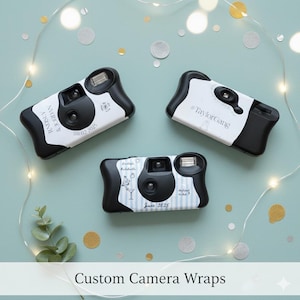 May include: Three black and white disposable cameras with custom wraps. The wraps feature names, dates, and text. The cameras are arranged on a light blue surface with decorative lights and confetti.