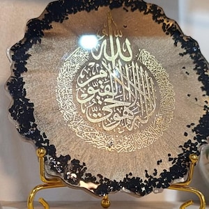 Puede incluir: Una placa de pared decorativa dorada y negra con caligrafía árabe. La placa está sobre un soporte de metal dorado.