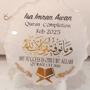 Op de afbeelding: Witte hars plaque met gouden belettering en een gouden Koran symbool. De plaque heeft de tekst "Isa Imran Awan Quran Completion Feb 2025" en "My success is only by Allah Quran (11:88)"