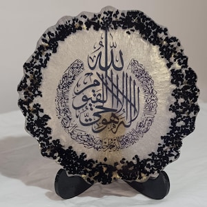 Può includere: Sottobicchiere rotondo in resina con calligrafia araba in inchiostro nero. Il sottobicchiere ha uno sfondo chiaro con dettagli dorati e un bordo nero strutturato. È esposto su un piccolo supporto nero.