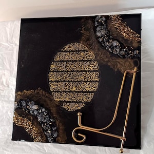 Op de afbeelding: Vierkant zwart kunstwerk met gouden Arabische kalligrafie. Het kunstwerk is versierd met gouden en zwarte decoratieve elementen. Een gouden metalen displaystandaard staat voor het kunstwerk.