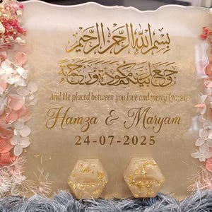 Könnte beinhalten: Dekoratives Hochzeitsschild mit goldener Schrift und floralen Akzenten. Das Schild trägt die Namen "Hamza & Maryam" und das Datum "24-07-2025". Es ist mit rosa und weißen Blumen und Blattgold verziert.