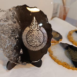 May include: A black and white resin coaster with a silver inscription of Arabic calligraphy. The coaster is on a black stand and has a gold rim.