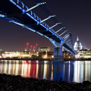May include: A modern pedestrian bridge with blue lights illuminating the underside, spanning a river at night. The bridge is lit up with blue lights, and the reflection of the lights can be seen in the water. The city skyline is visible in the background, including a large dome-shaped building.