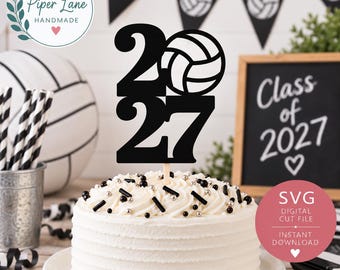 May include: A black cake topper featuring the numbers 2027 with a volleyball graphic integrated into the zero. The topper is displayed on a white frosted cake decorated with black and silver sprinkles, positioned in front of a chalkboard sign.