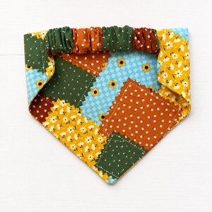 May include: A triangular pet bandana featuring a patchwork design of various floral and checkered fabrics in shades of mustard yellow, forest green, burnt orange, and light blue, with an elasticized gathered top edge for a secure fit.