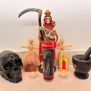 May include: A collection of spiritual items including a black skull, a red and black candle with a figure, a statue of a robed skeleton, two straw dolls, a mortar and pestle, a small card, and two lit candles.