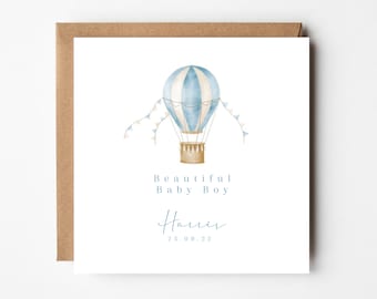 Puede incluir: Tarjeta de felicitación cuadrada con una ilustración en acuarela de un globo aerostático azul y blanco. El texto "BEAUTIFUL BABY BOY" está encima del nombre "Harris" y la fecha "25.09.22". La tarjeta está en un sobre marrón.