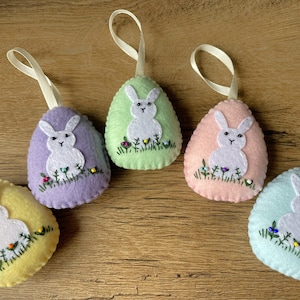 以下が含まれることがあります： 白いウサギが刺繍されたフェルト製のイースターエッグオーナメントが5つ。卵は、パープル、グリーン、ピンク、ライトブルー、イエローの異なる色です。各ウサギは、刺繍された目と小さなピンクの鼻を持っています。