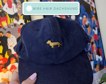 Puede incluir: Una gorra de béisbol de pana azul marino con un dachshund bordado en oro en la parte delantera. El texto "WIRE HAIR DACHSHUND" está impreso en una etiqueta azul claro.