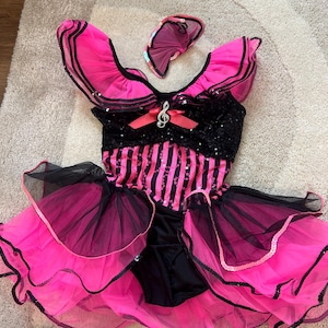 May include: A vibrant pink and black dance costume with a matching headpiece. The bodice features black sequins and a pink bow with a musical note charm. The skirt has layers of pink tulle and black trim. A fun and eye-catching outfit.