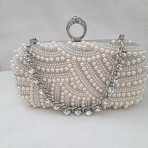 May include: An ivory-coloured, oval clutch bag covered in small, round pearls. A silver chain with clear, sparkling accents drapes across the front. The top features a ring-shaped handle encrusted with rhinestones. A glamorous evening bag.