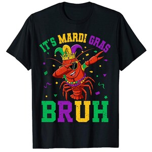 May include: Black t-shirt with a green, purple, and yellow graphic of a cartoon crawfish wearing a jester hat and sunglasses. The text "It's Mardi Gras bruh" is printed on the shirt.