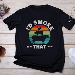 May include: Black t-shirt with a graphic design featuring a cow, pig, and chicken stacked on top of each other, with the text "I'd Smoke That" in white. The design is set against a colorful sunset background.