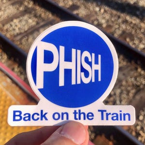 May include: A white sticker with a blue circle containing the word "PHISH" in white letters. Below the circle, the text "Back on the Train" is printed in blue. The sticker is held up against a blurred background of train tracks.