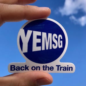 May include: A white sticker with a blue circle containing the letters "YEMSG". Below the circle, the text "Back on the Train" is printed in blue. The sticker is held against a blue sky with clouds.