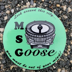 May include: A round, green sticker with the text "Look around this city... M S GOOSE you must be out of your mind!" in black. The sticker features a black and white illustration of a stadium.