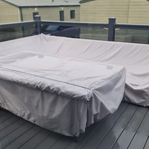 May include: A gray patio furniture set covered with white protective covers. The set includes a table and a couch. The furniture is on a gray wooden deck.