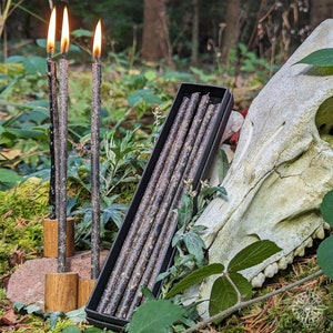 Könnte beinhalten: Eine Schachtel mit schwarzen Kerzen in Kegelform mit einer Holzstruktur. Die Kerzen sind in einer Schachtel mit schwarzem Deckel angeordnet. Die Schachtel liegt auf einem weißen Schädel.