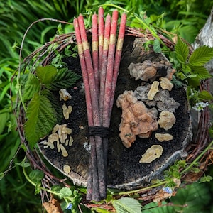 May include: A bundle of red and gold incense sticks tied together with a black string, resting on a wooden surface with small pieces of amber resin scattered around.
