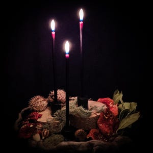Könnte beinhalten: Drei schwarze Kerzenständer mit brennenden Kerzen, umgeben von getrockneten Pflanzen und Moos. Die Kerzenflammen haben eine weiße Mitte mit roten und blauen Akzenten. Die Anordnung ist vor einem dunklen Hintergrund.