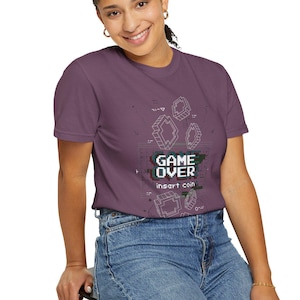 Puede incluir: Una camiseta morada con las palabras "GAME OVER" e "insert coin" en una fuente pixelada. La camiseta también presenta formas y líneas geométricas blancas. La persona lleva vaqueros azules.