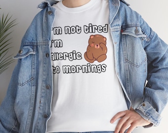 Op de afbeelding: Wit T-shirt met een bruine cartoonbeer en de tekst "Ik ben niet moe, ik ben allergisch voor ochtenden".