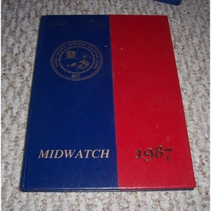 Puede incluir: Un libro de tapa dura azul y rojo con el texto "MIDWATCH 1987" en la portada. El libro tiene un sello dorado con el texto "NAVAL RESERVE OFFICERS TRAINING CORPS MIT" en la portada.