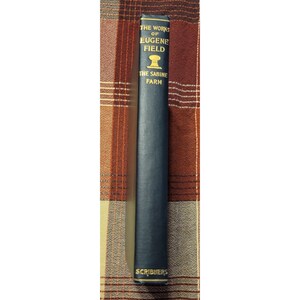 Puede incluir: The Works of Eugene Field, The Sabine Farm, un libro negro con letras doradas en el lomo. El libro está sobre una superficie a cuadros marrón y beige.