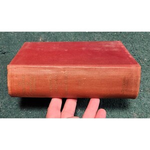 Puede incluir: Un libro antiguo con una cubierta roja descolorida. El lomo muestra texto en relieve, incluido el título y el autor. Los bordes del libro muestran signos de desgaste, lo que sugiere edad y uso. El libro está sostenido por una mano.