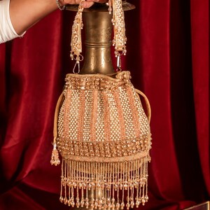 May include: A gold and beige drawstring handbag featuring vertical beaded stripes, a decorative band of metallic sequins, and a long fringe of dangling beads at the base. The bag includes a matching beaded handle and a drawstring closure mechanism.