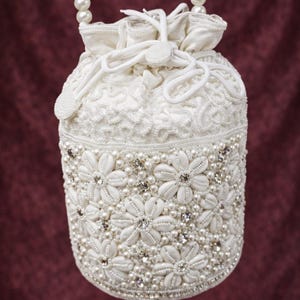 May include: A white beaded drawstring bag with a pearl handle. The cylindrical bag features a floral design with pearls and rhinestones. The top is gathered with a drawstring closure. The handle is made of white pearls.