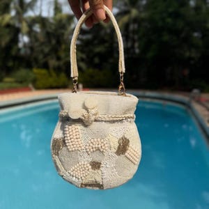 Può includere: Una borsetta color crema con perline, di forma arrotondata e con manico. La borsa presenta una varietà di dimensioni e motivi di perline, con accenti dorati. Il manico è di colore crema abbinato.