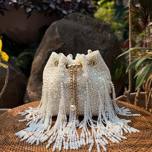 Puede incluir: Un bolso blanco con cuentas y cordón, con una cadena dorada y detalles de perlas. El bolso está cubierto de pequeñas cuentas blancas y tiene largas borlas de cuentas. El bolso está sobre una superficie tejida.
