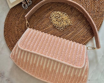 Könnte beinhalten: Eine blushrosa Perlen-Clutch mit passendem Schulterriemen und goldener Kette. Die rechteckige Tasche hat vertikale Reihen aus rosa Perlen und weißen Streifen. Goldene Creolen und ein Buch sind im Hintergrund zu sehen.