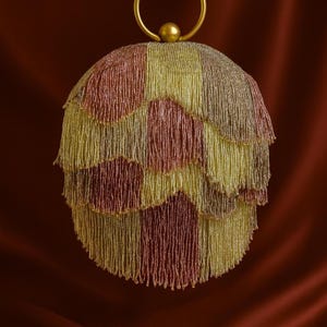 Puede incluir: Un bolso de mano con cuentas y asa dorada. El bolso esférico presenta un diseño en capas con secciones alternas de cuentas doradas, rosas y burdeos. El asa es un simple anillo dorado sujeto a la parte superior del bolso.