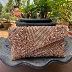 May include: A blush-toned beaded clutch bag with a fold-over flap. The bag has a geometric design with rows of beads and sequins, and is embellished with clear and pearl-like beads. A stylish accessory for special occasions.