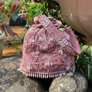 Könnte beinhalten: Eine staubrosa Handtasche mit aufwendiger Blumenstickerei und Perlenbesatz. Die Tasche hat einen Kordelzugverschluss, einen Schulterriemen mit Perlen und eine Fransenborte am unteren Rand. Das Design ist mit Pailletten und kleinen, weißen Perlen verziert.