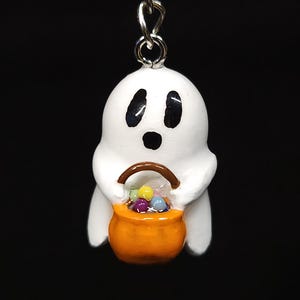 May include: A white ghost-shaped keychain charm with black eyes and mouth, holding an orange trick-or-treat bucket filled with colorful candies. The charm is attached to a silver-colored clasp and chain, perfect for Halloween.